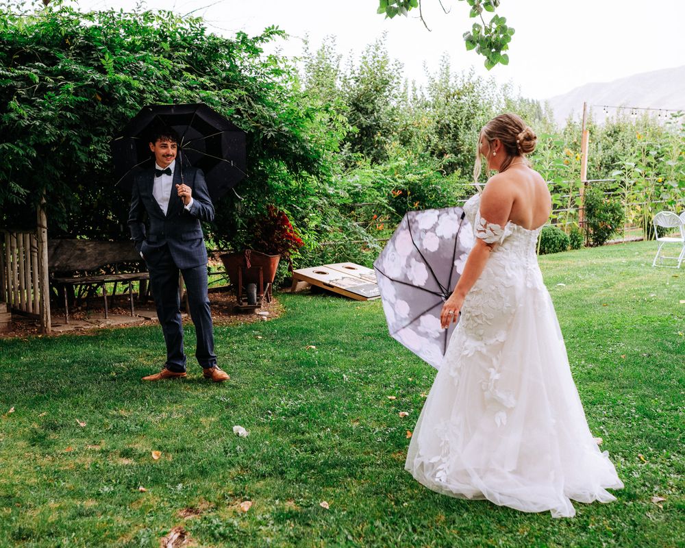 Bride in white lace gown and groom in navy suit share a rainy outdoor wedding moment with umbrellas in a lush garden.