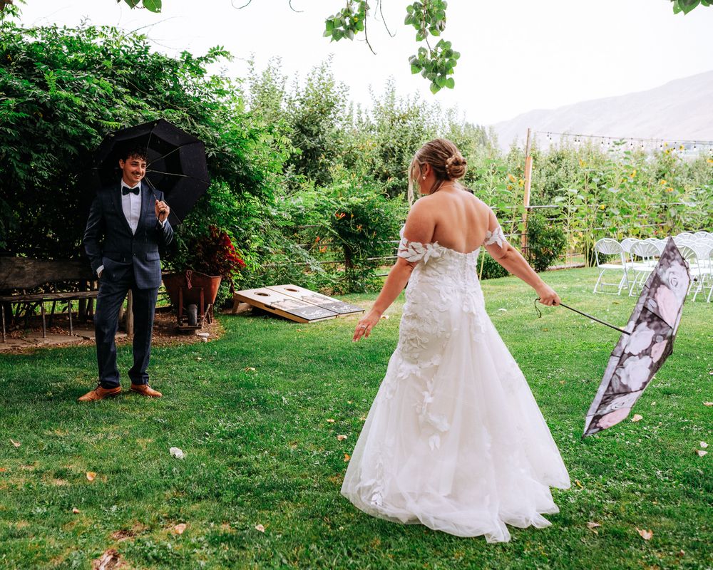 Bride in a lace gown approaches groom holding an umbrella during an outdoor garden wedding ceremony.
