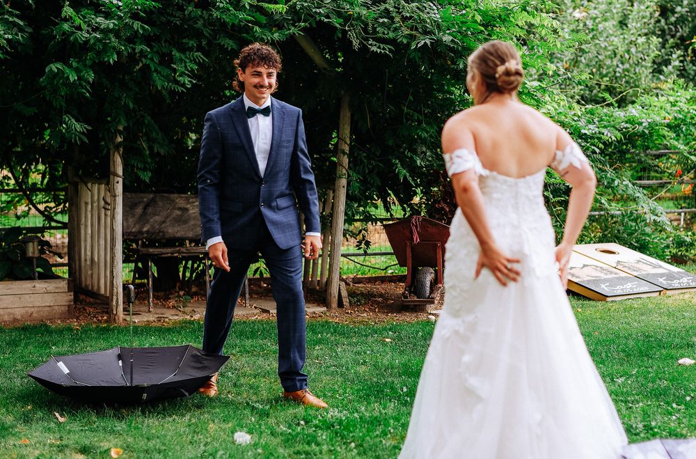 Groom in navy tuxedo smiling at bride in white off-shoulder wedding dress during outdoor first look ceremony.