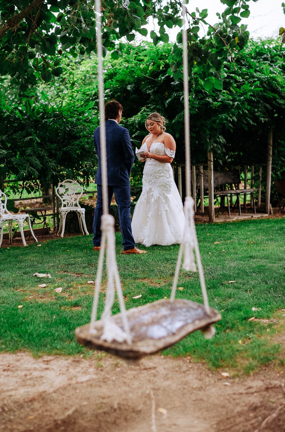 Bride in lace wedding gown reading vows to groom outdoors, with rustic wooden swing in foreground.