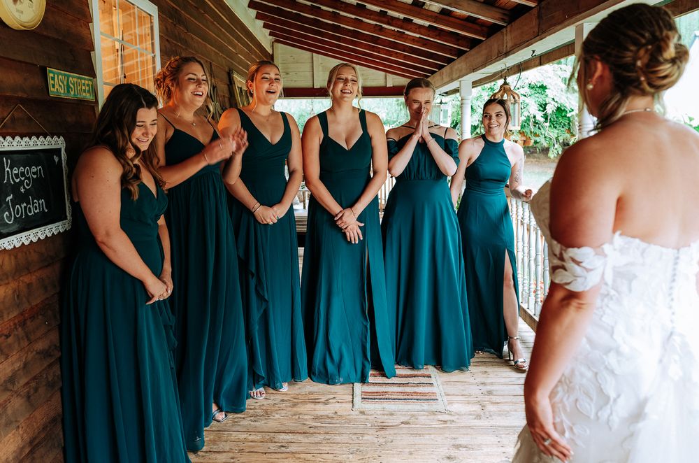 Six bridesmaids in teal gowns react emotionally during first look with bride on rustic wooden porch.