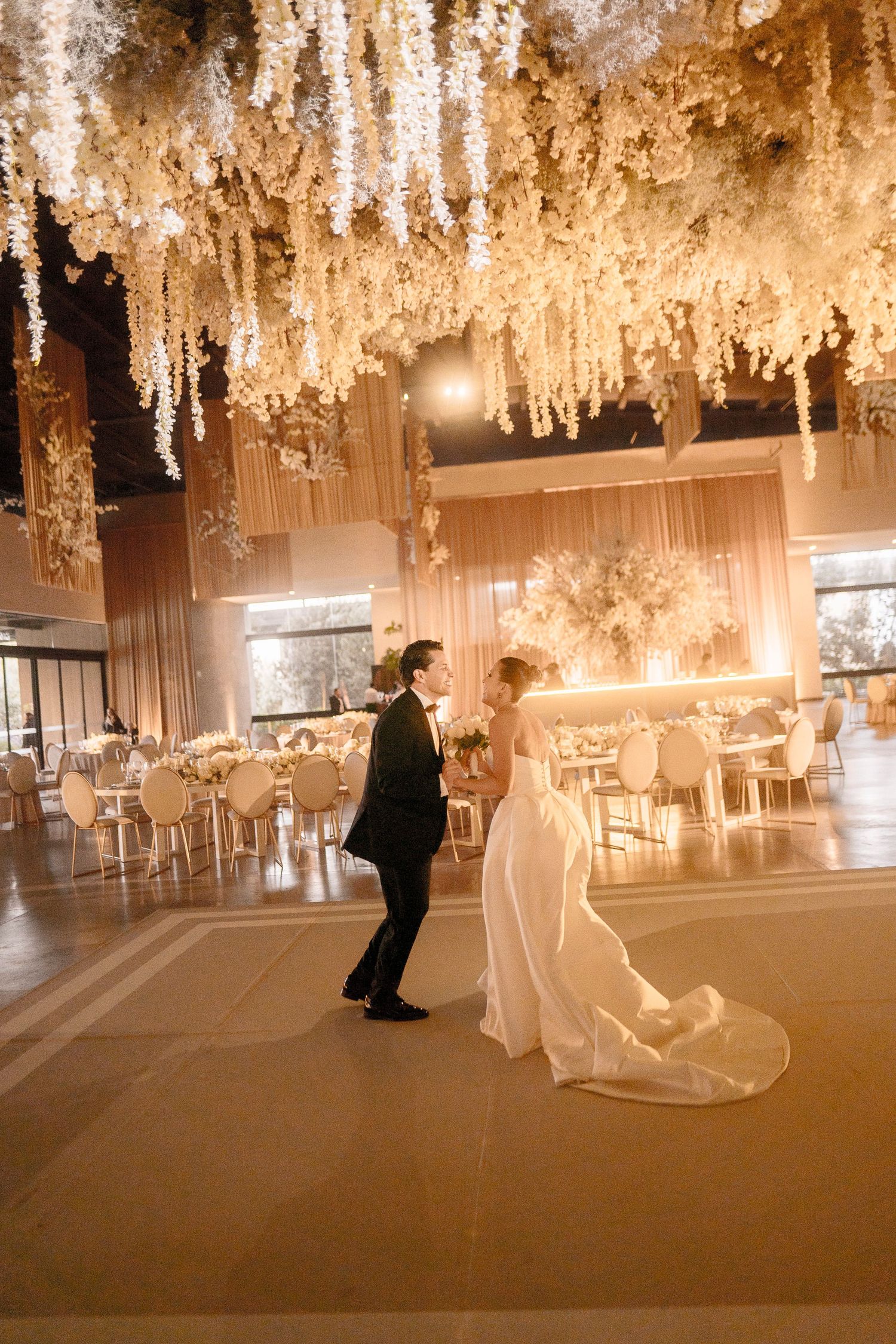 Bride and groom share a romantic dance under stunning white floral ceiling installation in an elegant wedding reception venue.