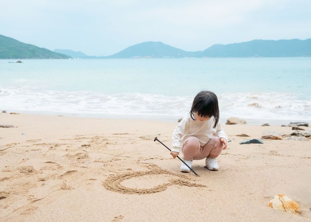 A 2-year-old toddler drawing in the sand with a stick during a candid beach family photography session in Hong Kong