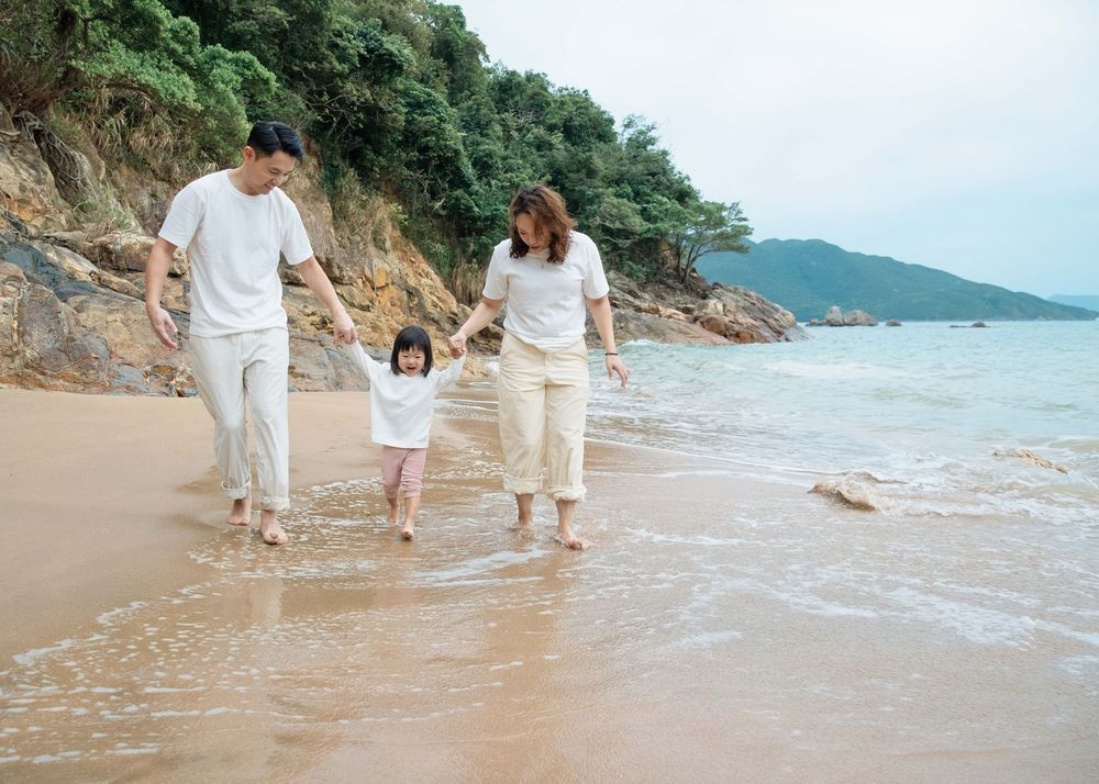 A family of three walking together on a quiet sandy beach with rocky cliffs and ocean views during a Hong Kong family photo shoot