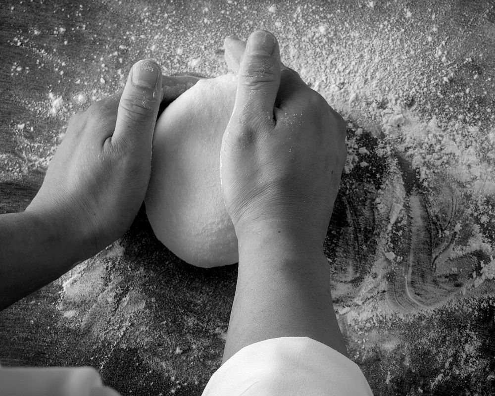 Hands kneading dough on a flour-dusted surface in a black and white close-up photo.