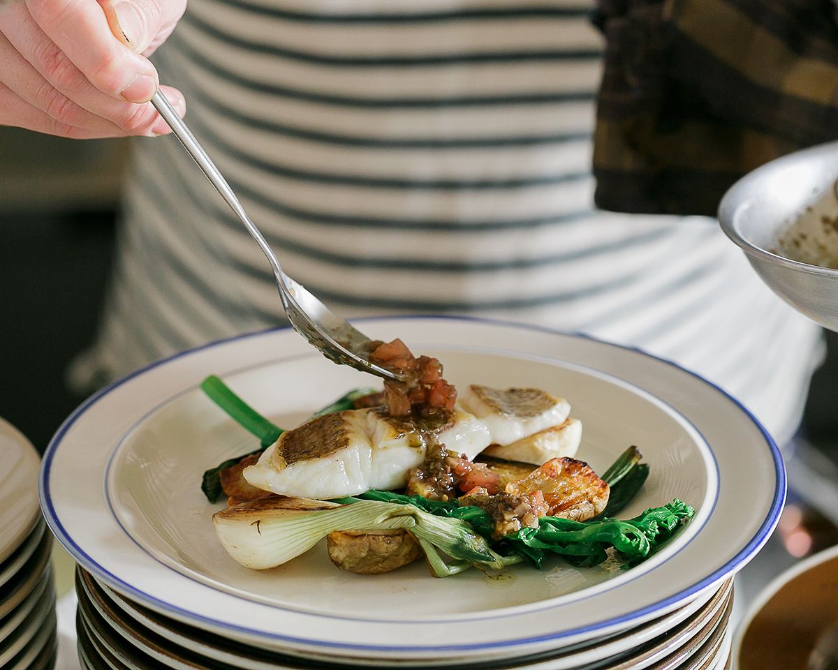 A hand holding a fork over a plate of grilled fish with bok choy and tomato sauce, stacked on white plates.