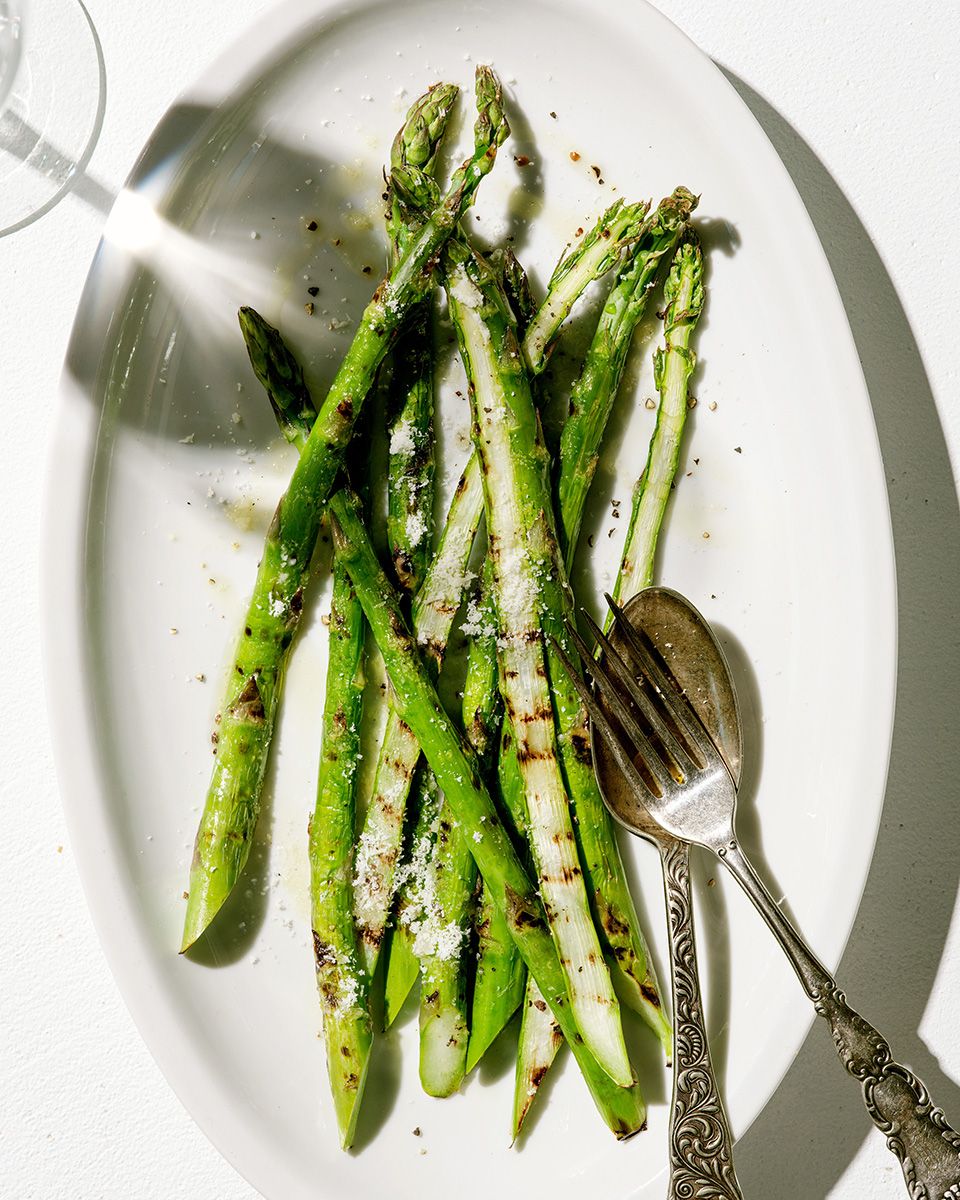 Grilled asparagus spears topped with Parmesan and black pepper on a white oval plate with silver fork and spoon.