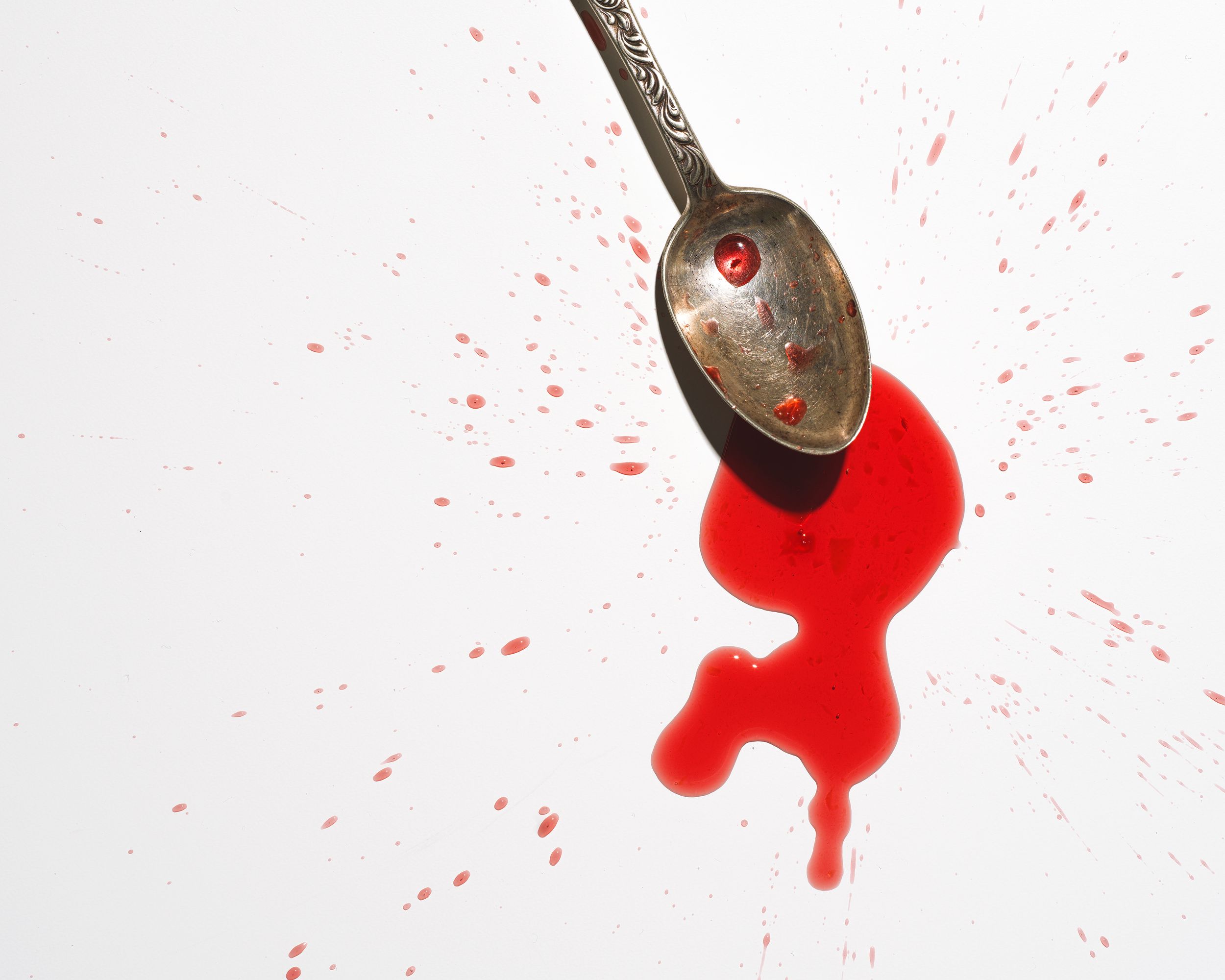 A silver spoon with red liquid splattered on a white background, resembling a crime scene or food photography.