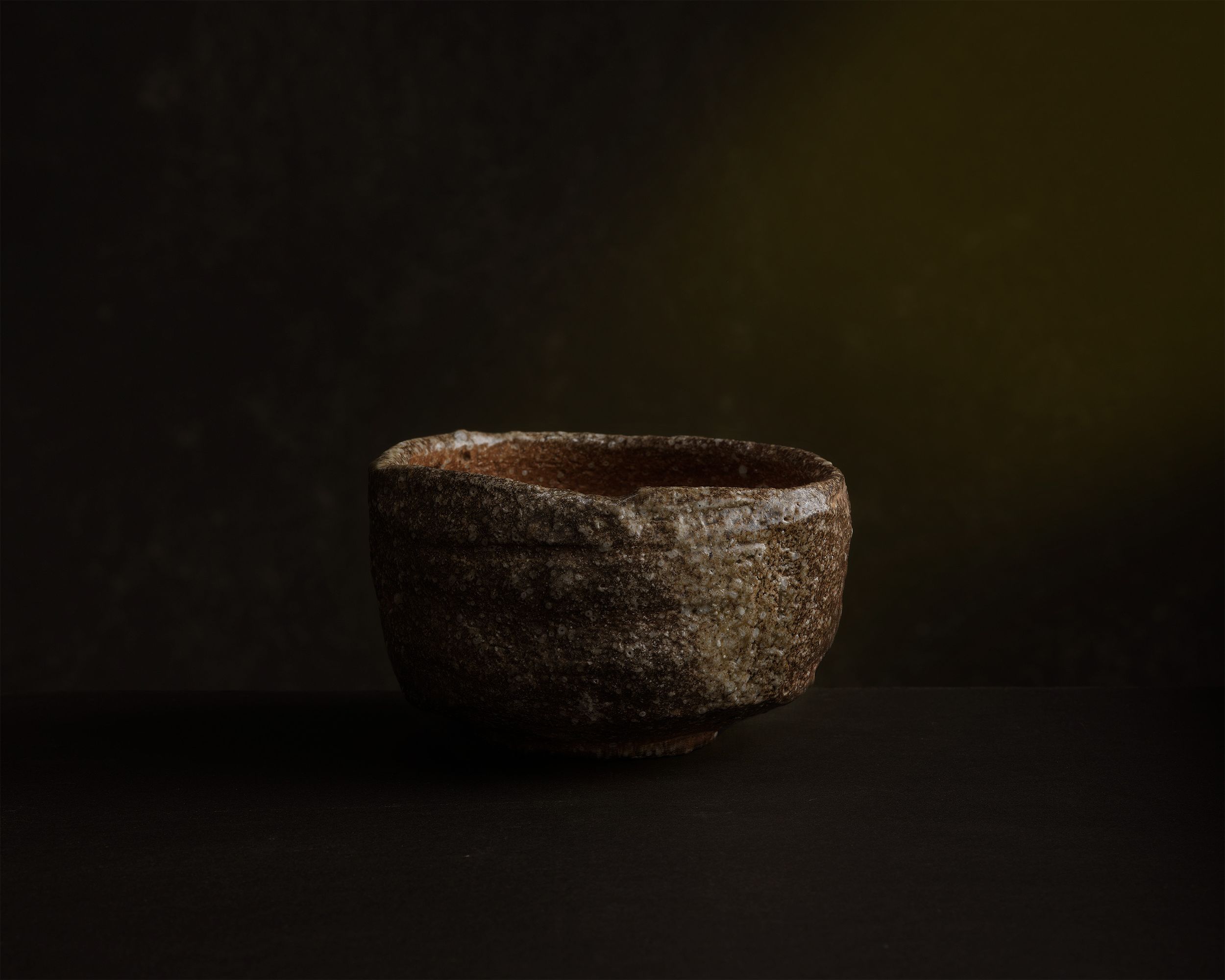 Rustic handmade dark ceramic tea bowl with rough textured surface on a dark background.