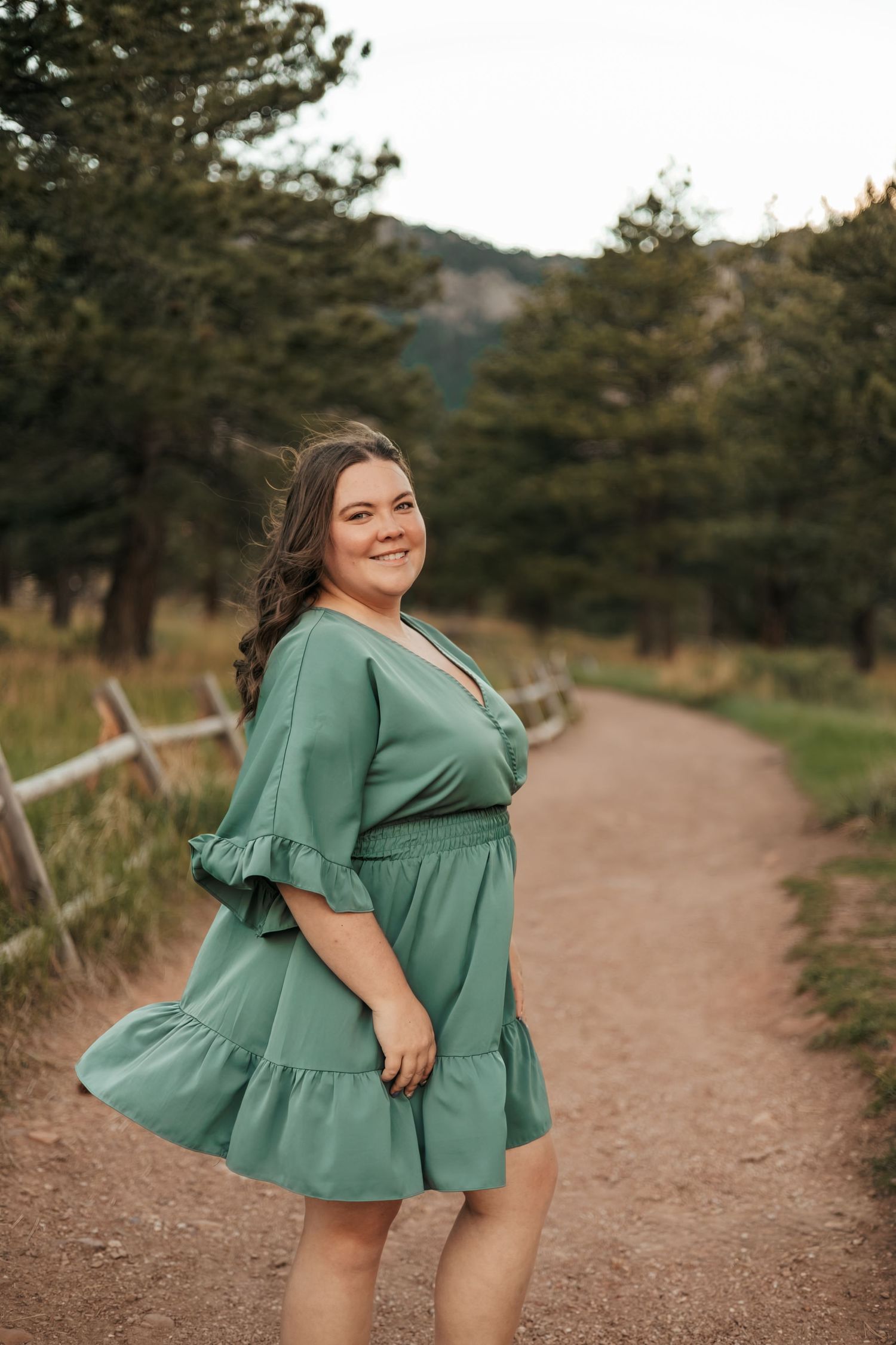 Woman in teal dress smiling on a scenic dirt path surrounded by trees and mountains | Baby Announcement Photographer in Boulder, Colorado