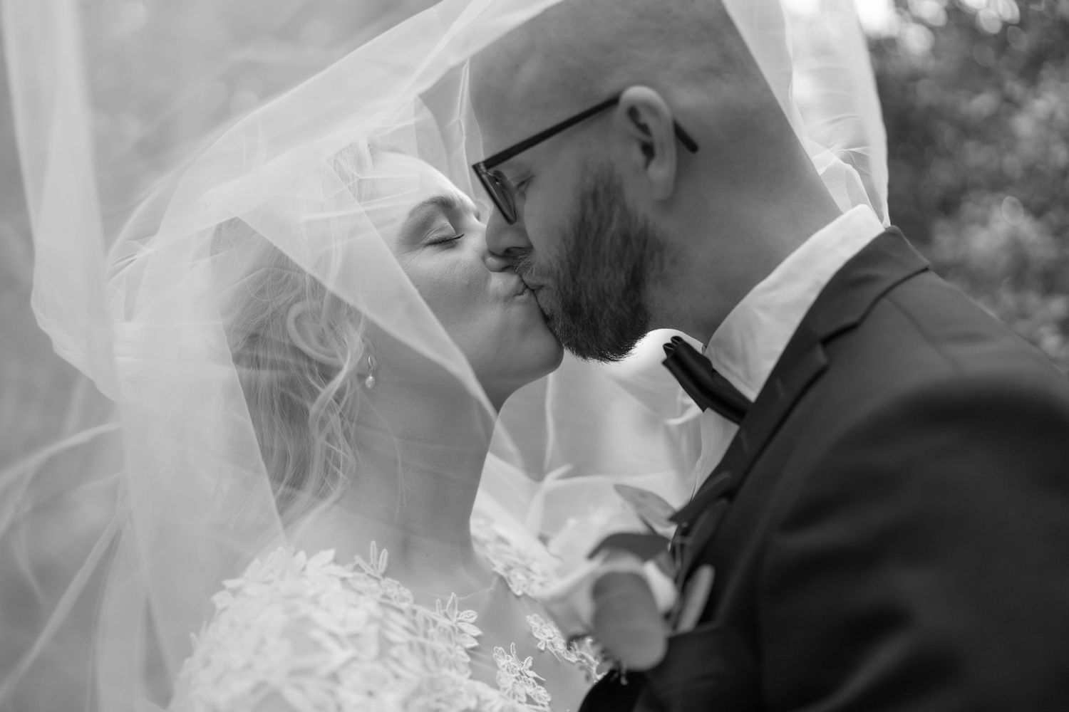Black and white photo of a bride and groom sharing a romantic kiss under a flowing wedding veil.