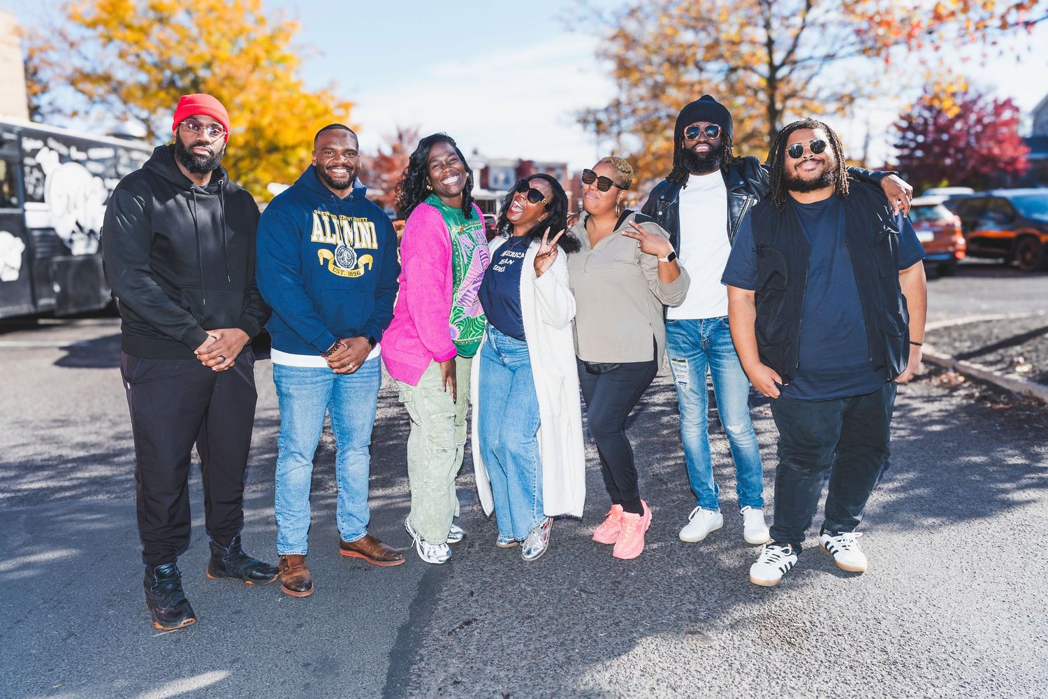 Group of seven friends posing together outdoors on a sunny autumn day with colorful fall foliage in the background.