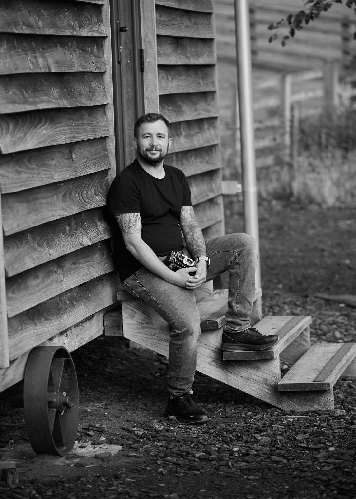 Tattooed man in black t-shirt holding camera, leaning against wooden building on steps, black and white portrait photo.