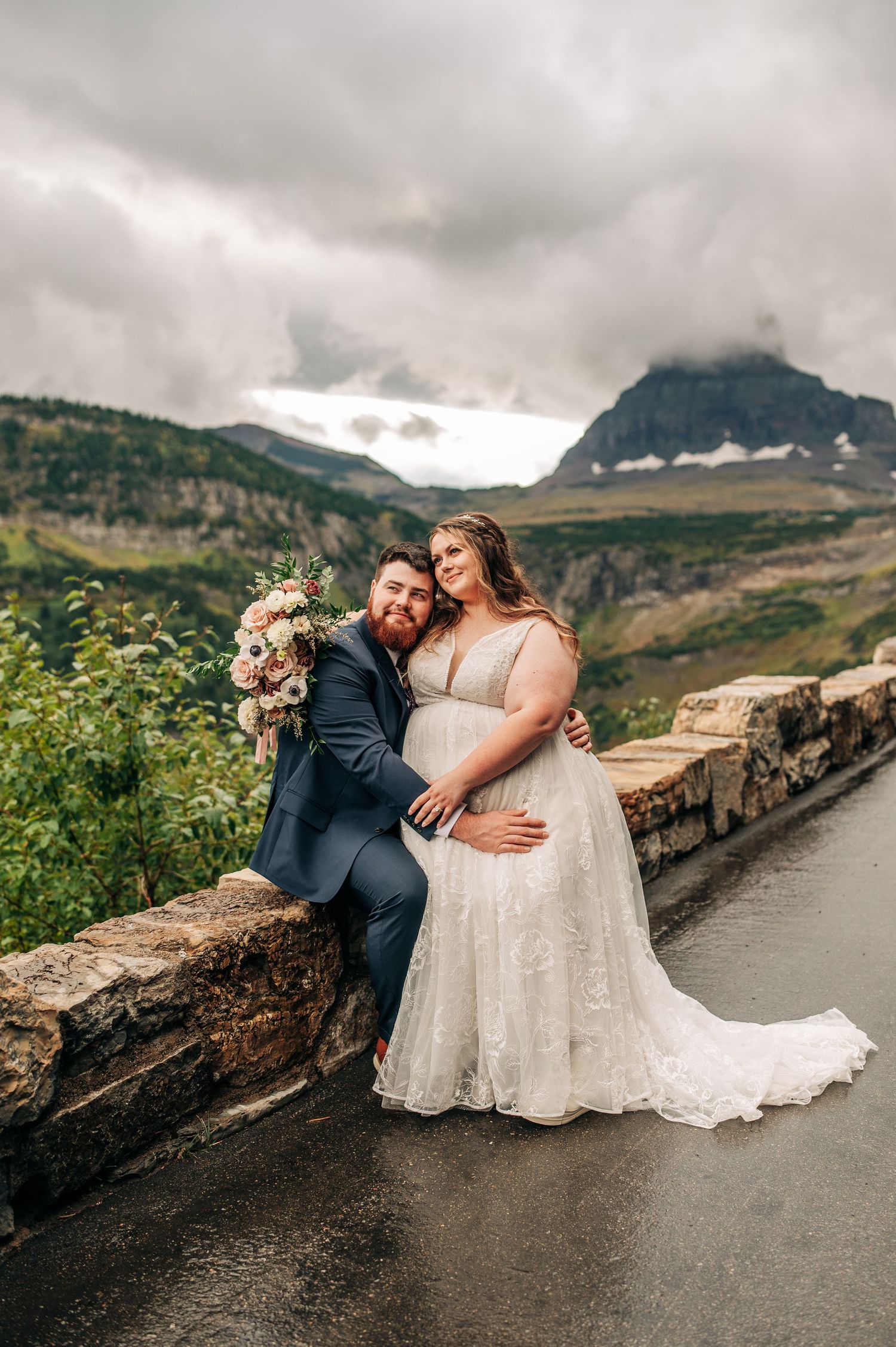 Bride and groom pose on a stone wall with dramatic mountain scenery and cloudy skies at Glacier National Park.