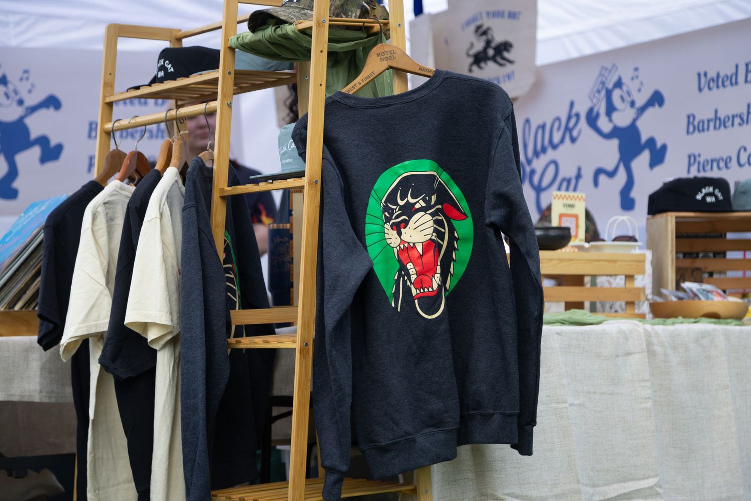 Black sweatshirt with panther graphic displayed on wooden rack at outdoor market vendor booth.