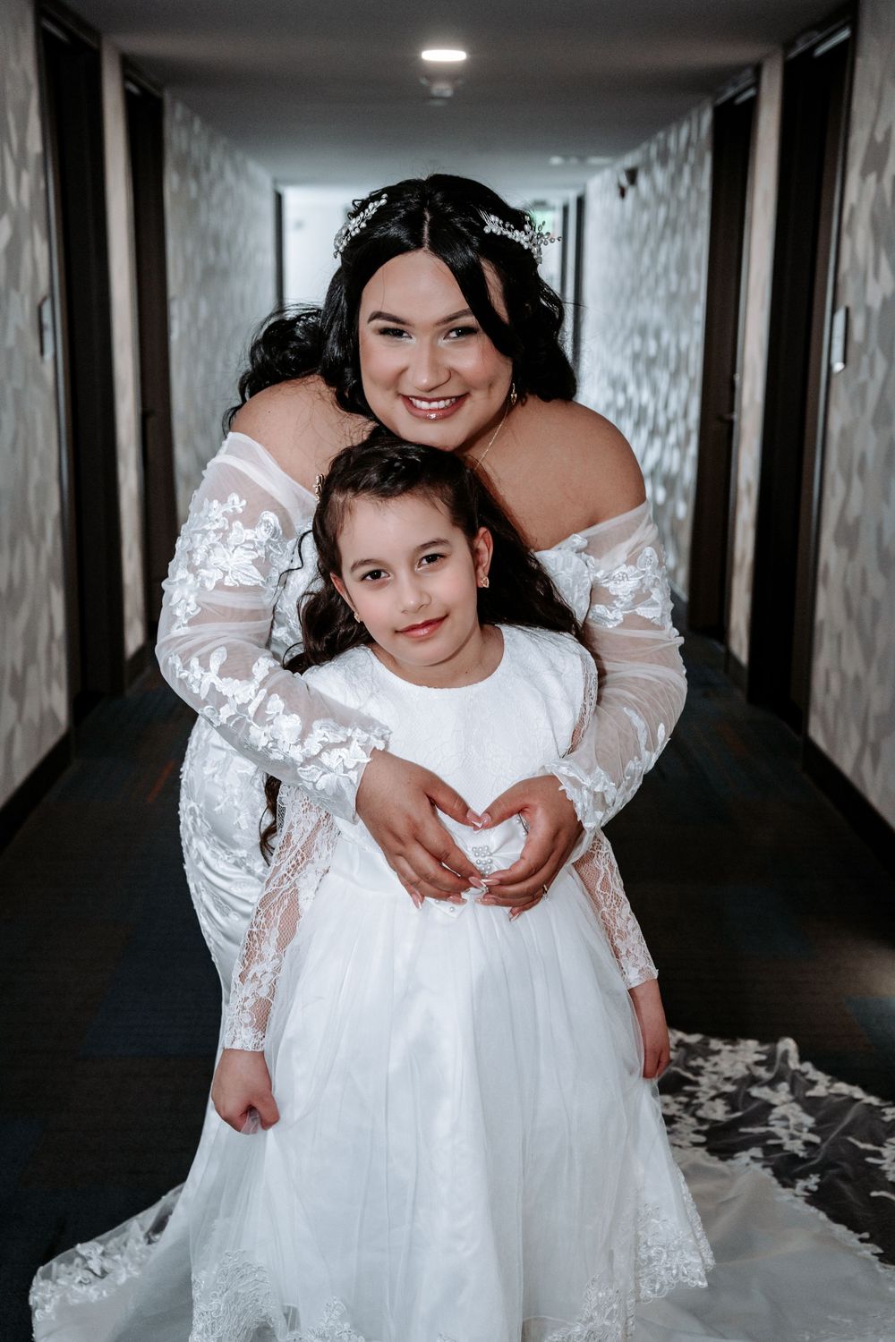 Bride in lace wedding gown hugs flower girl in white dress, making heart shape with hands in hotel hallway.