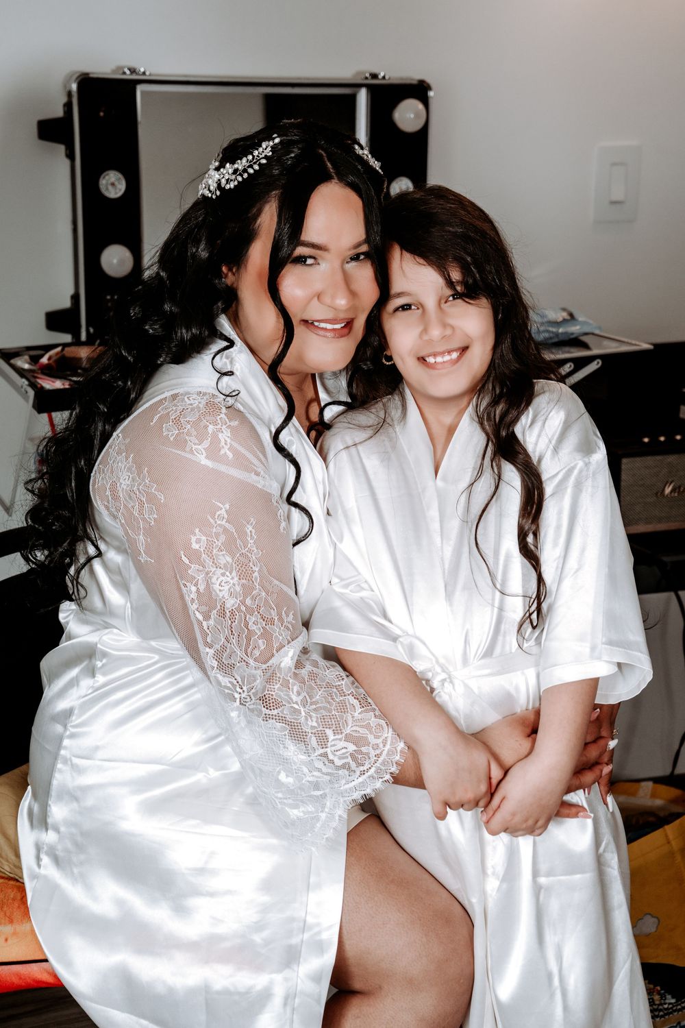 Bride in white lace robe hugs smiling young girl in satin robe during wedding preparation.