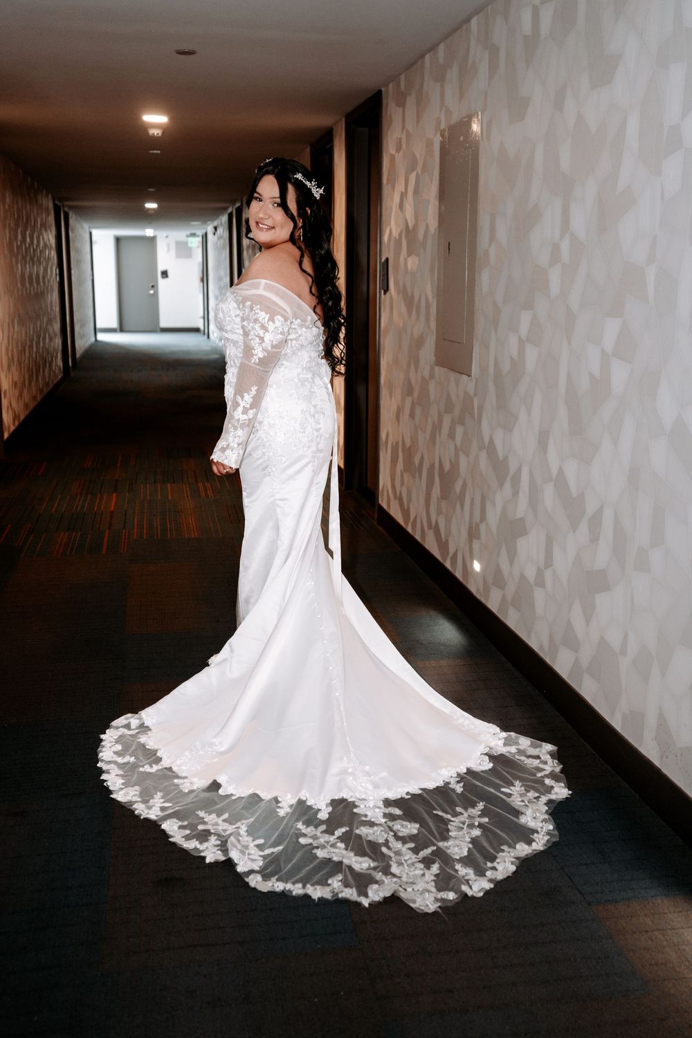 Bride in elegant white lace wedding gown with long train poses in hotel corridor, showcasing off-shoulder dress design.
