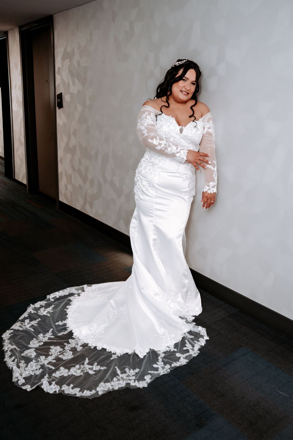 Bride in white lace mermaid wedding gown with long sleeves and cathedral train, posing elegantly in a hallway.