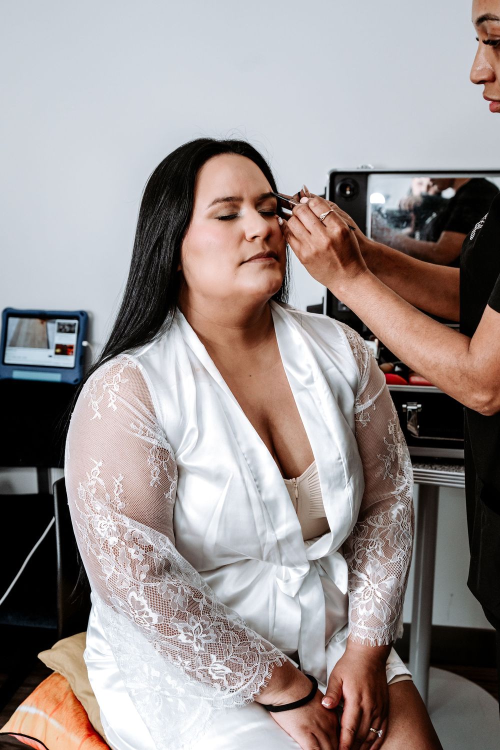 Makeup artist applies eyeshadow to bride wearing white lace robe during bridal preparation.