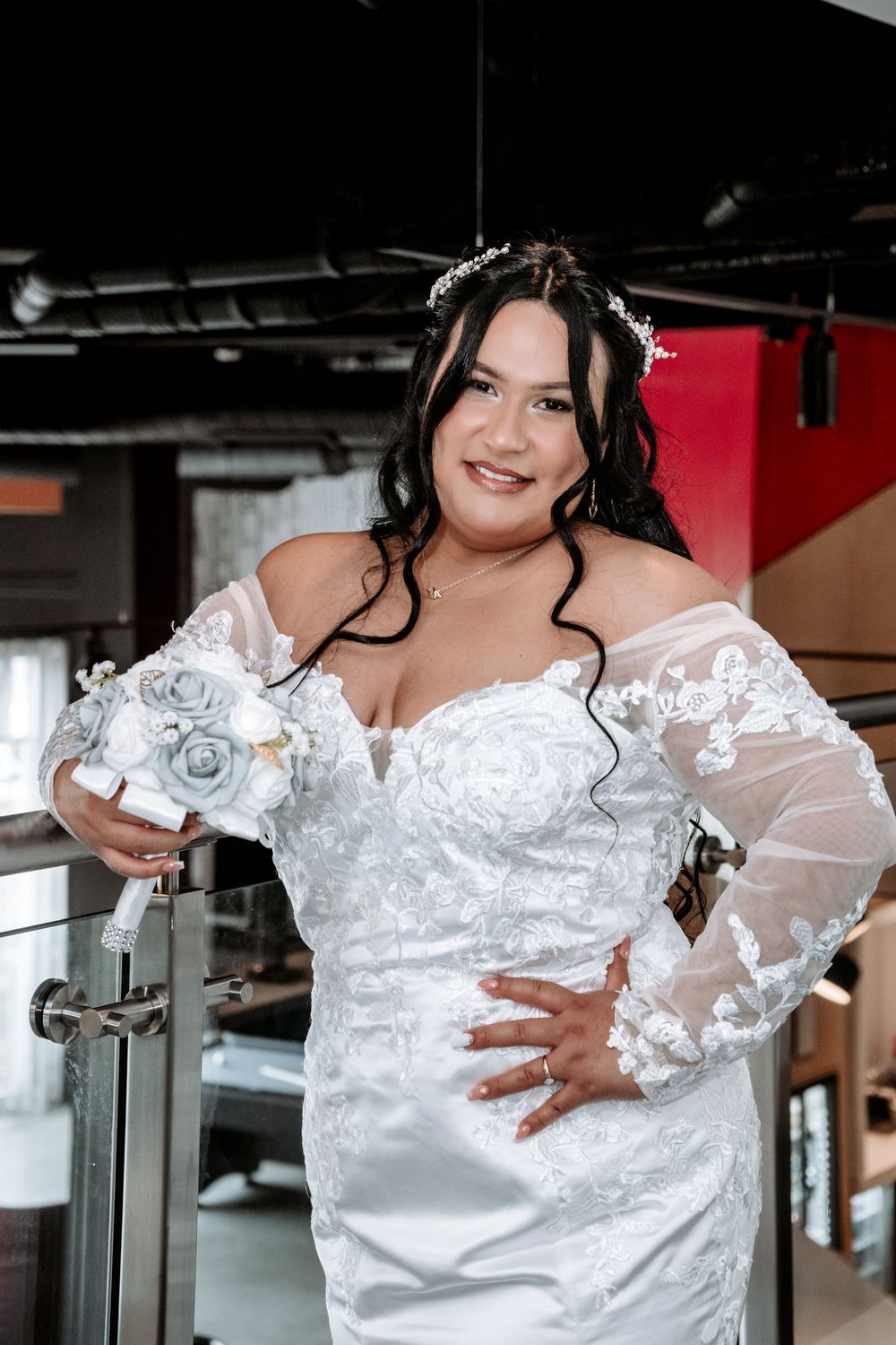 Plus-size bride in off-shoulder lace wedding gown holding gray rose bouquet posing elegantly indoors.