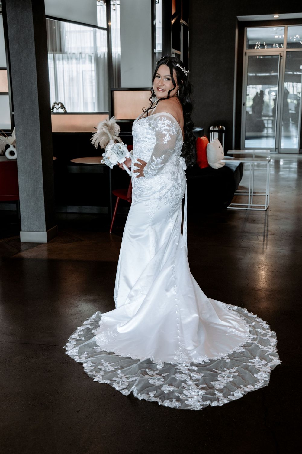 Bride in off-shoulder lace wedding gown with long train poses elegantly in venue interior.