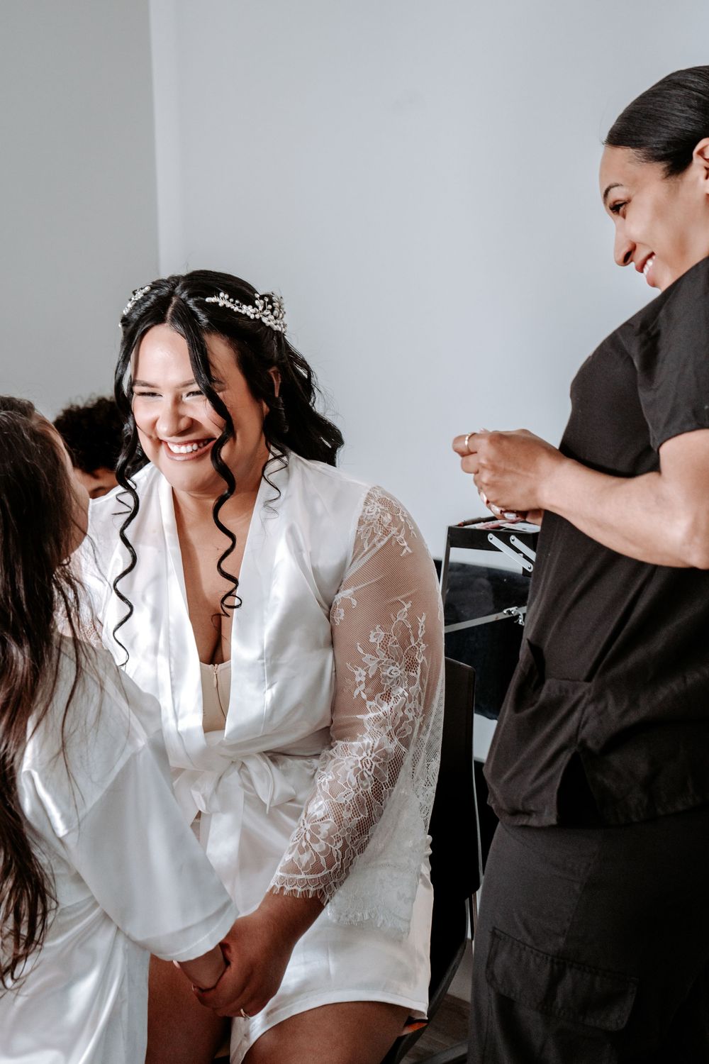 Bride in white lace robe and tiara laughing joyfully during wedding preparations with stylist nearby.
