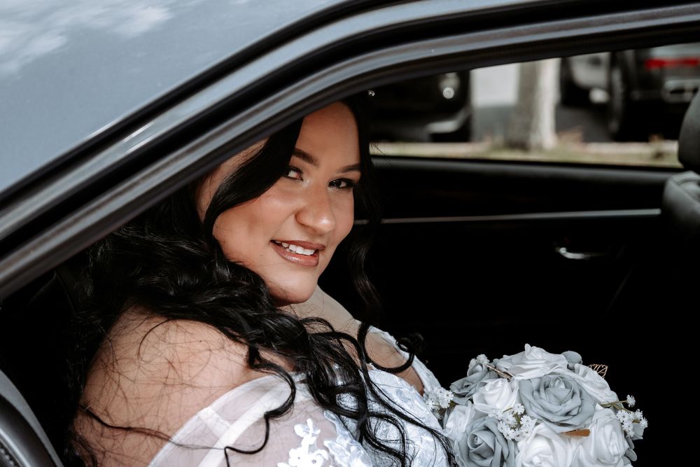 Smiling bride in white lace dress holding blue bouquet sitting in car window on wedding day.