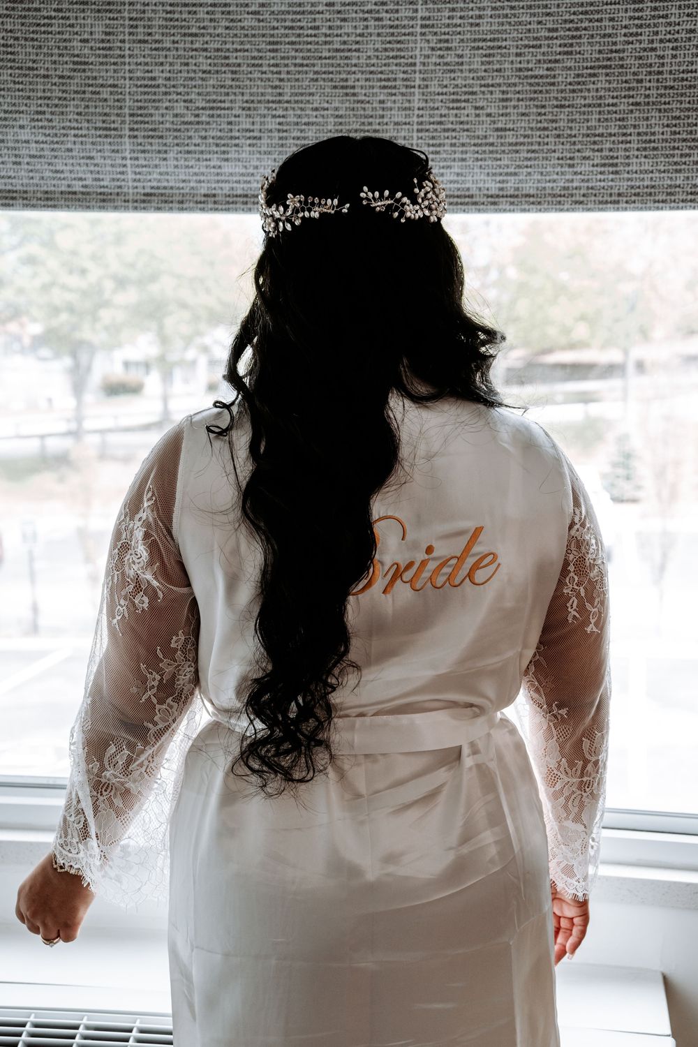 Bride in white lace robe with gold script stands backlit by window wearing floral hair crown.