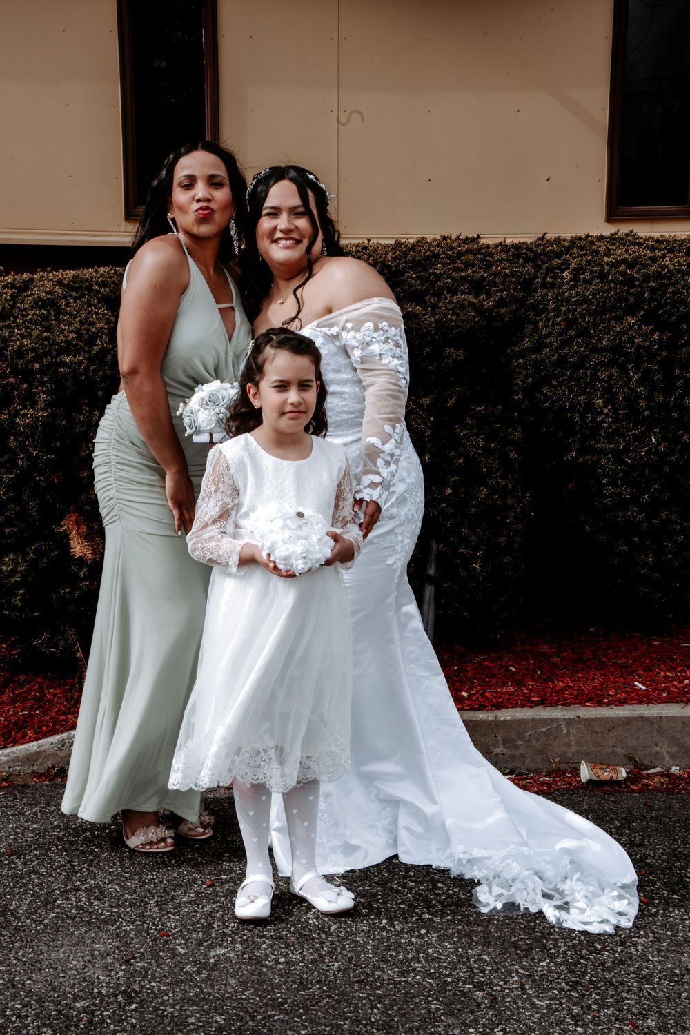 Bride in white gown poses with bridesmaid in sage green dress and flower girl in white dress outdoors.
