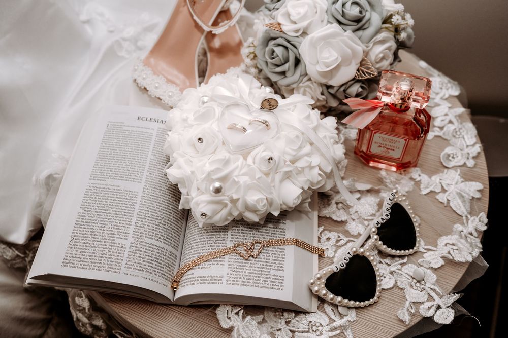 Wedding flat lay with white rose bouquet, open Bible, pearl necklace, sunglasses, and floral perfume bottle on lace.