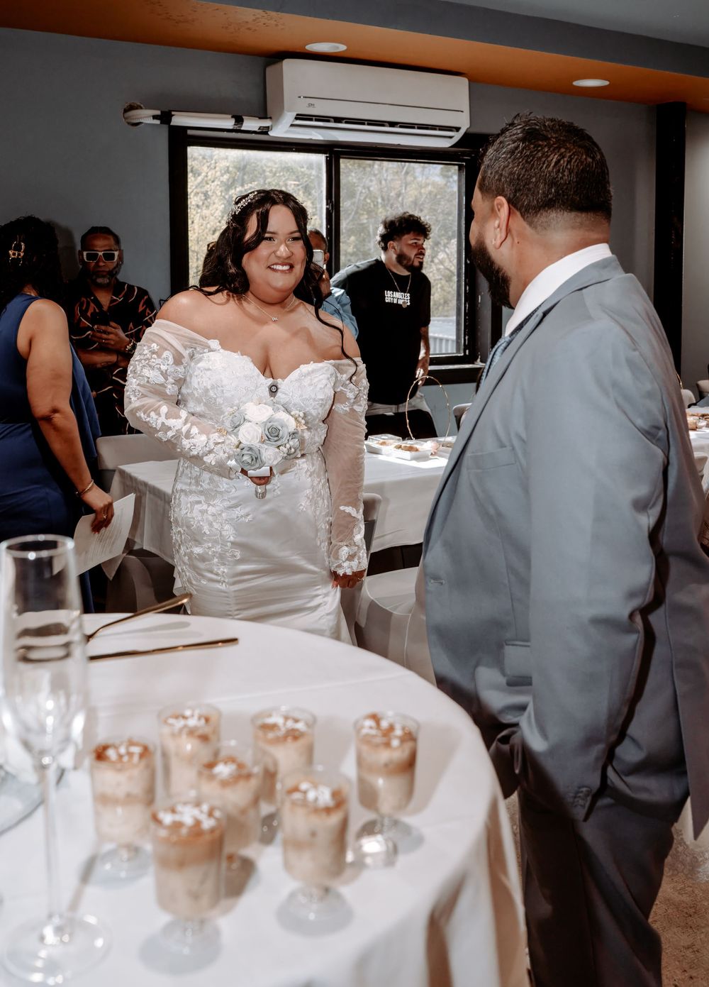 Bride in white off-shoulder gown smiles at groom in gray suit at wedding reception with dessert table.