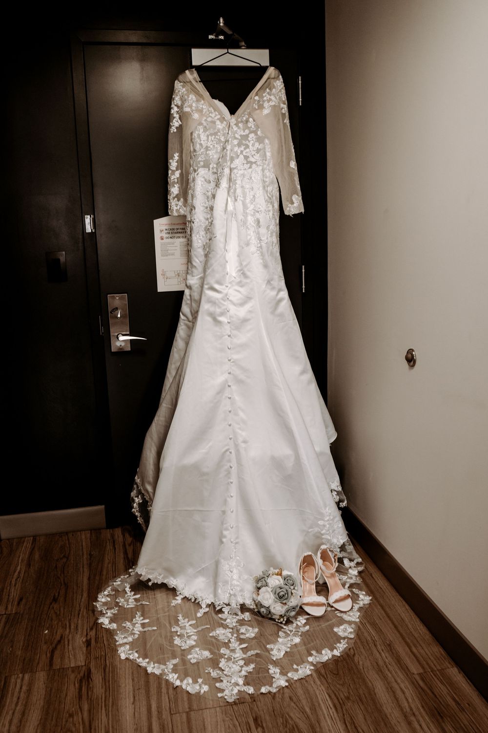 White lace wedding dress with long sleeves hanging on door with bridal bouquet and heels below.