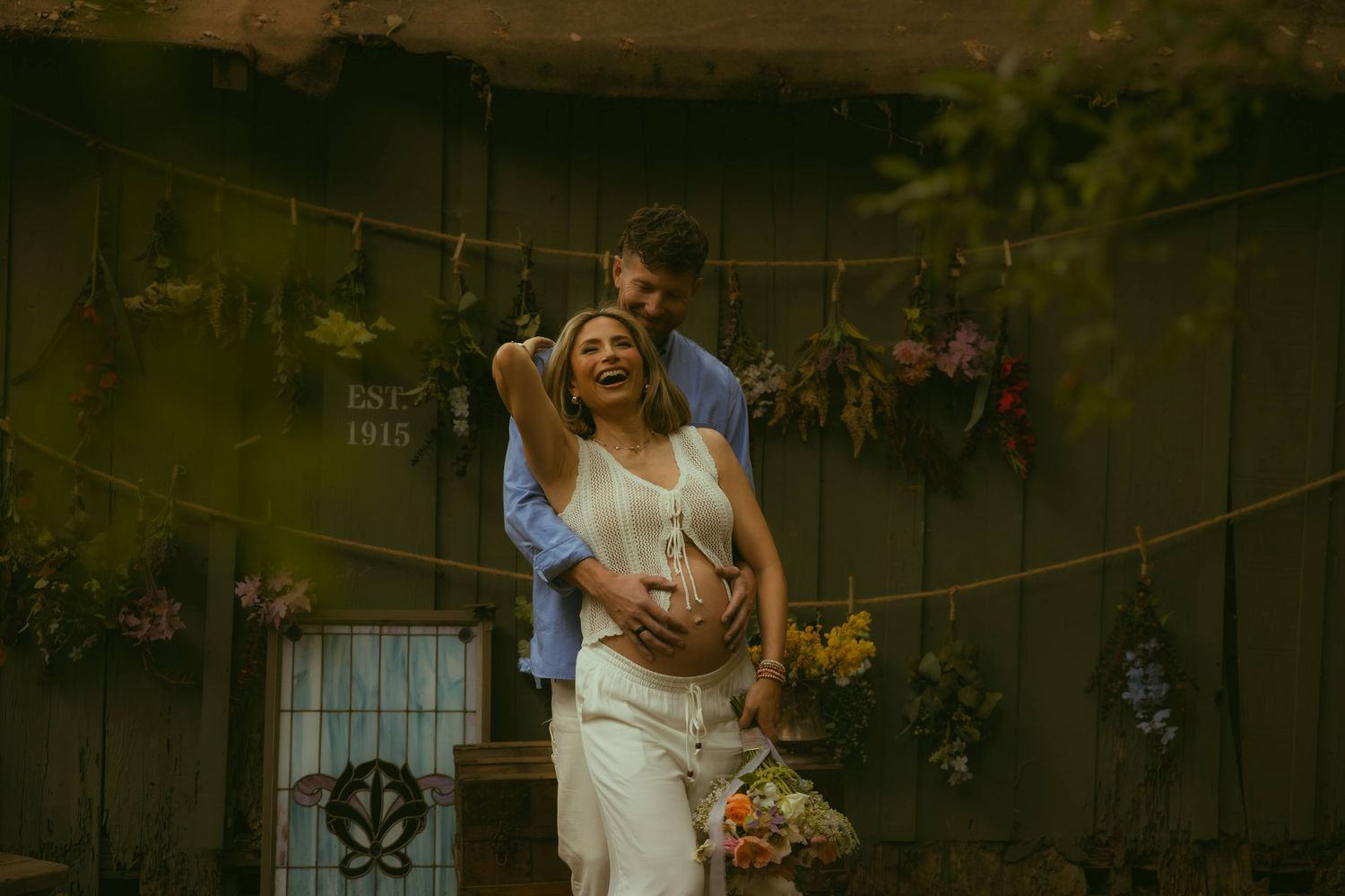 Couple embracing outdoors near a rustic wall with hanging dried flowers, woman in white dress holding a colorful bouquet.