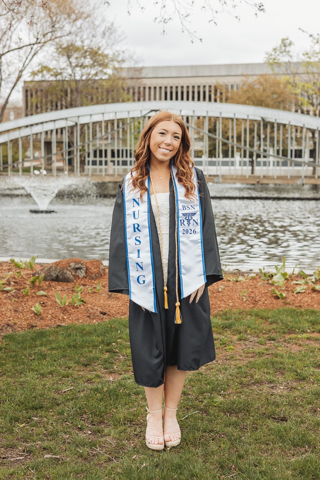 Smiling nursing graduate in black cap and gown with BSN 2026 stole stands near a white bridge and fountain.