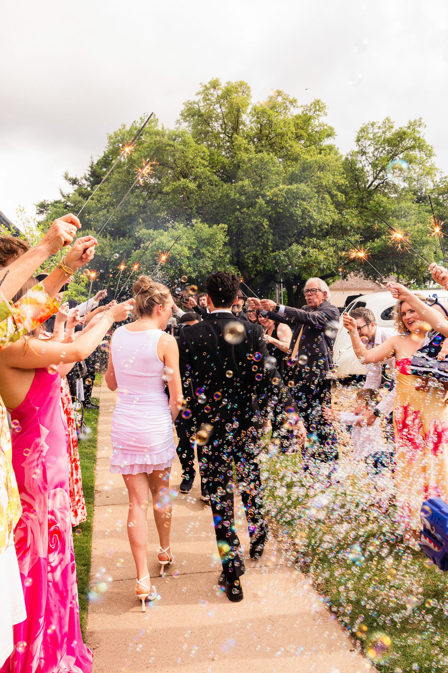 Wedding couple walks through a joyful confetti and sparkler sendoff surrounded by colorful guests outdoors.