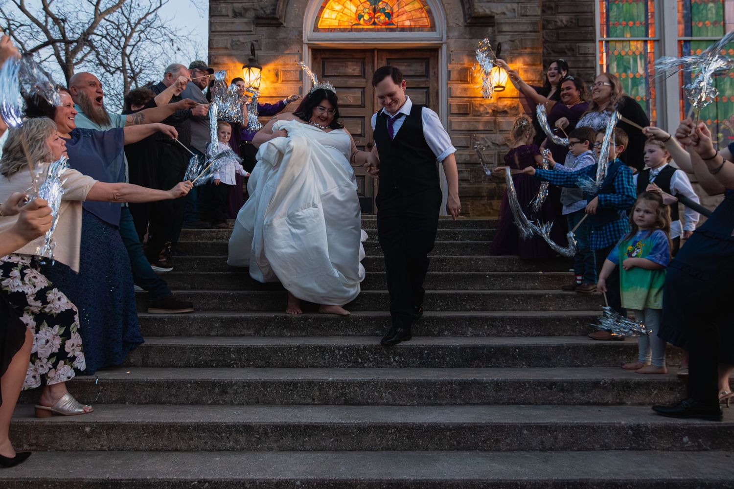 Newlywed couple exits stone church on steps lined with cheering guests at dusk wedding celebration.