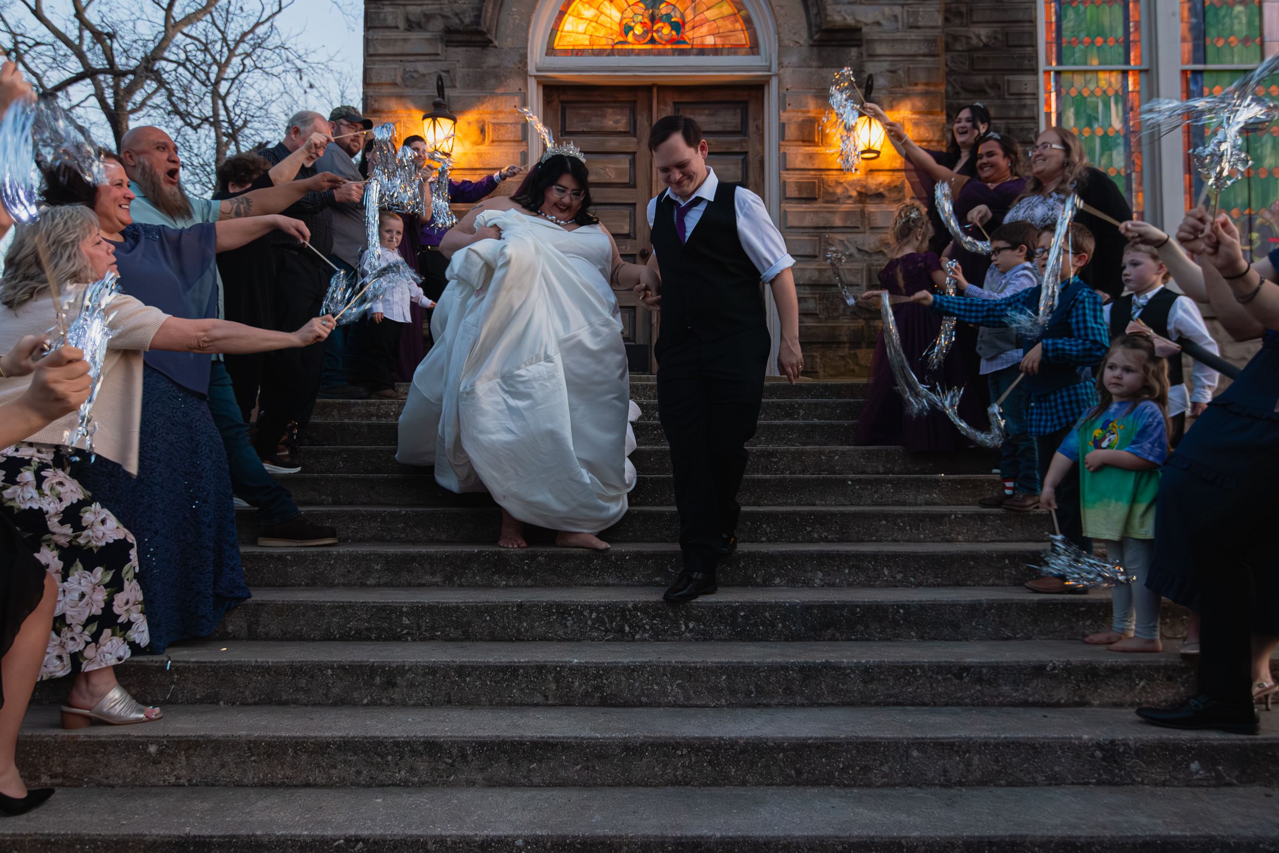 Newlywed couple exits stone church on steps lined with cheering guests at dusk wedding celebration.