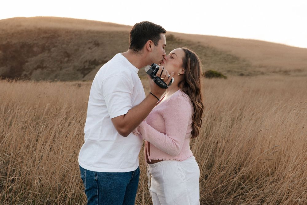 Couple sharing a romantic kiss in a golden grass field with rolling hills in the background.