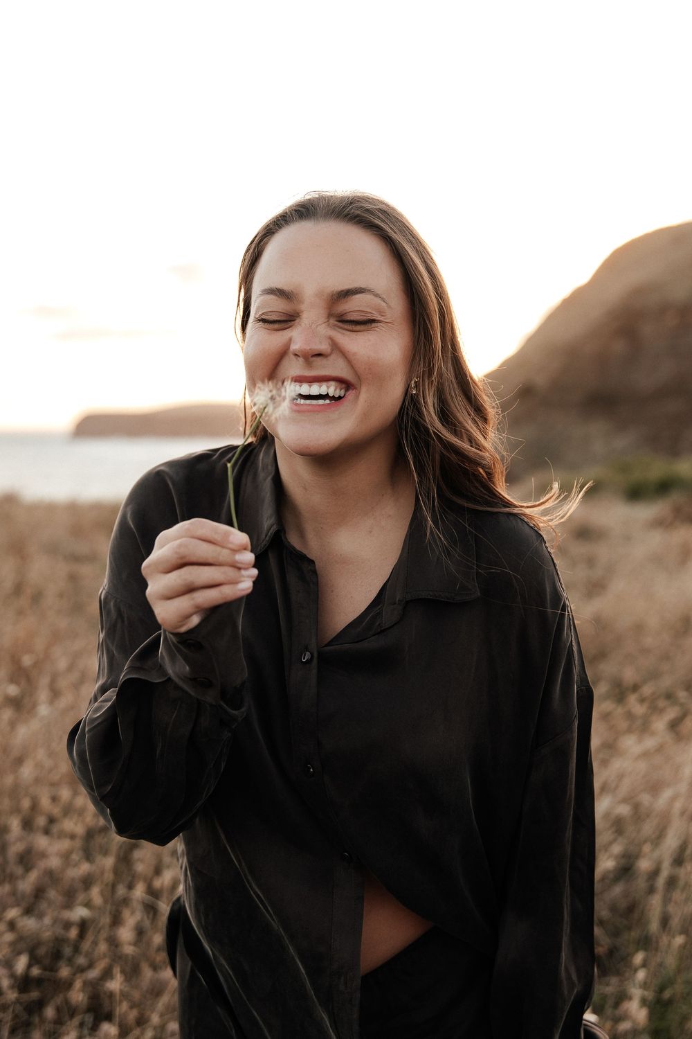 A joyful woman laughing outdoors holding a dandelion, wearing a black shirt with coastal cliffs at sunset.