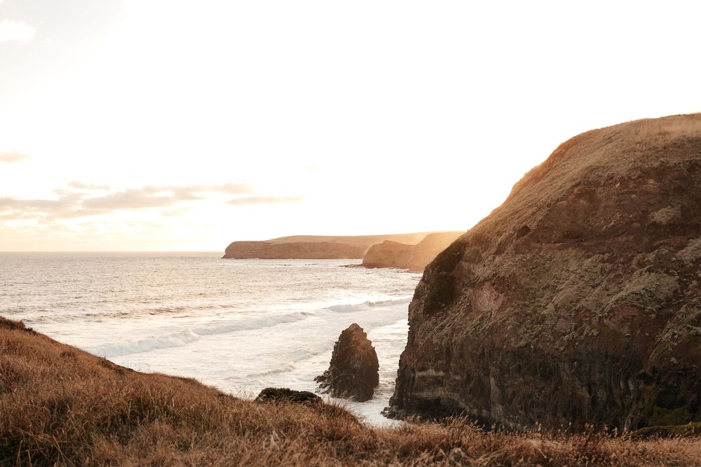 Golden hour coastal cliffs with rocky sea stacks and calm ocean waves under a soft glowing sunset sky.