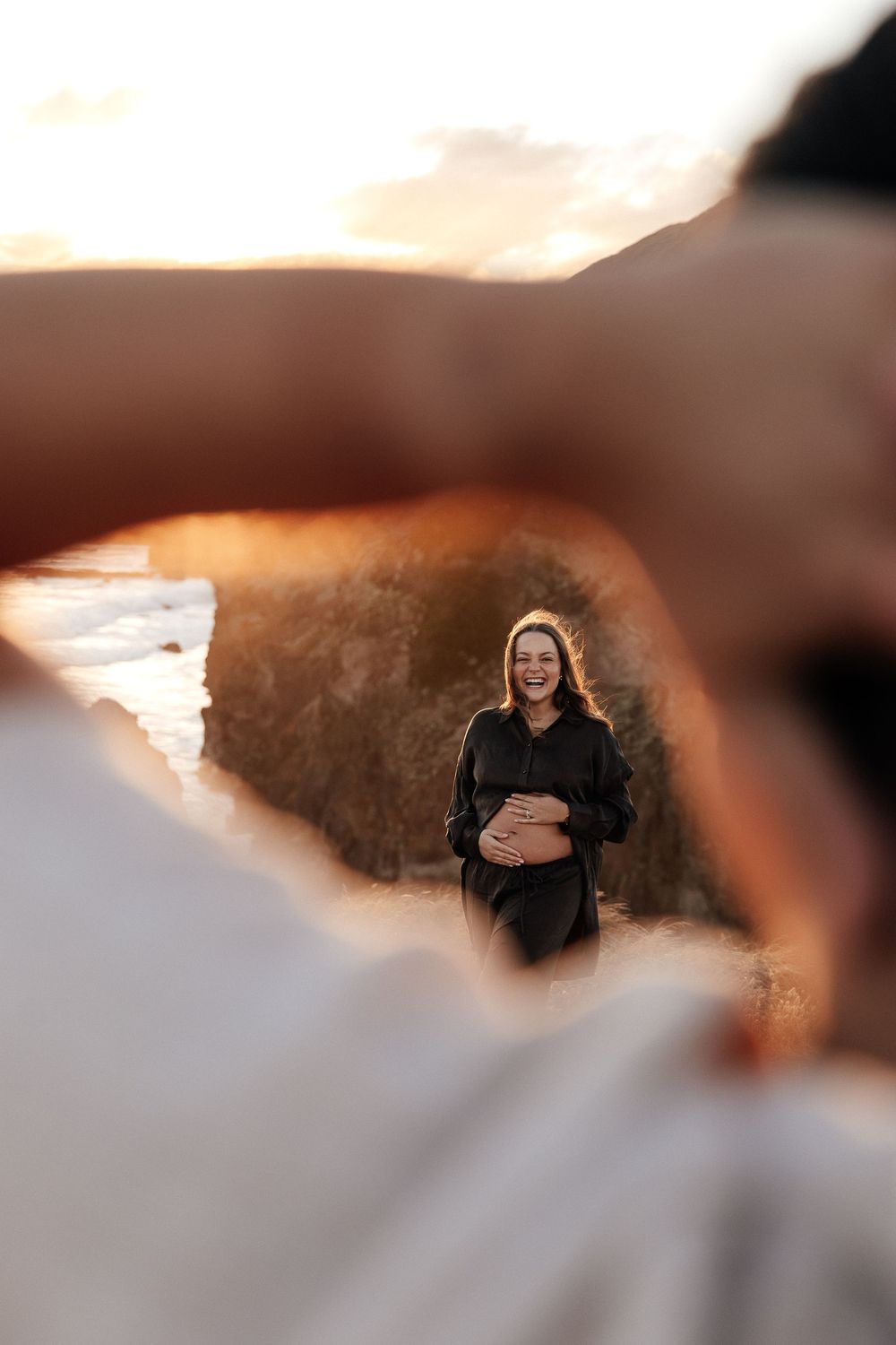 Pregnant woman in black dress smiling at sunset beach, framed by partner's hands in golden hour light.