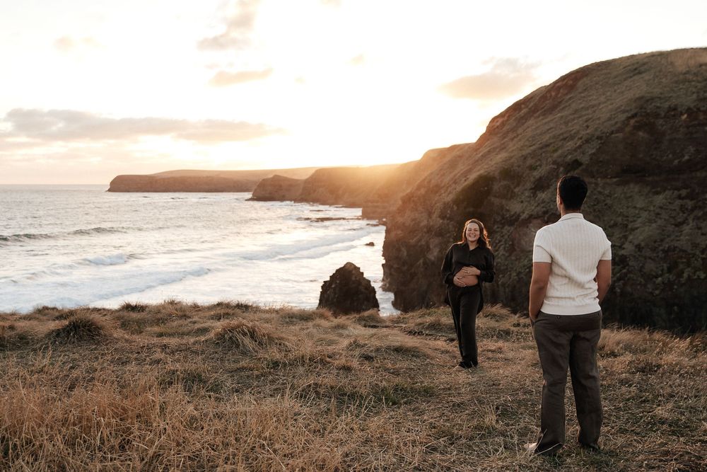 Couple standing on coastal cliffs at golden sunset with ocean waves below.