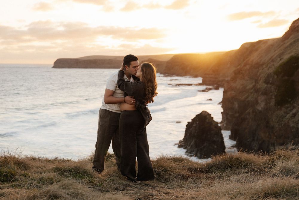 Couple embracing at golden sunset on coastal cliffs with ocean waves and rocky shoreline in background.