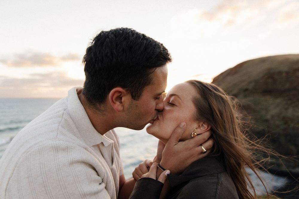 Couple sharing a romantic kiss on a rocky beach at sunset with ocean waves in the background.