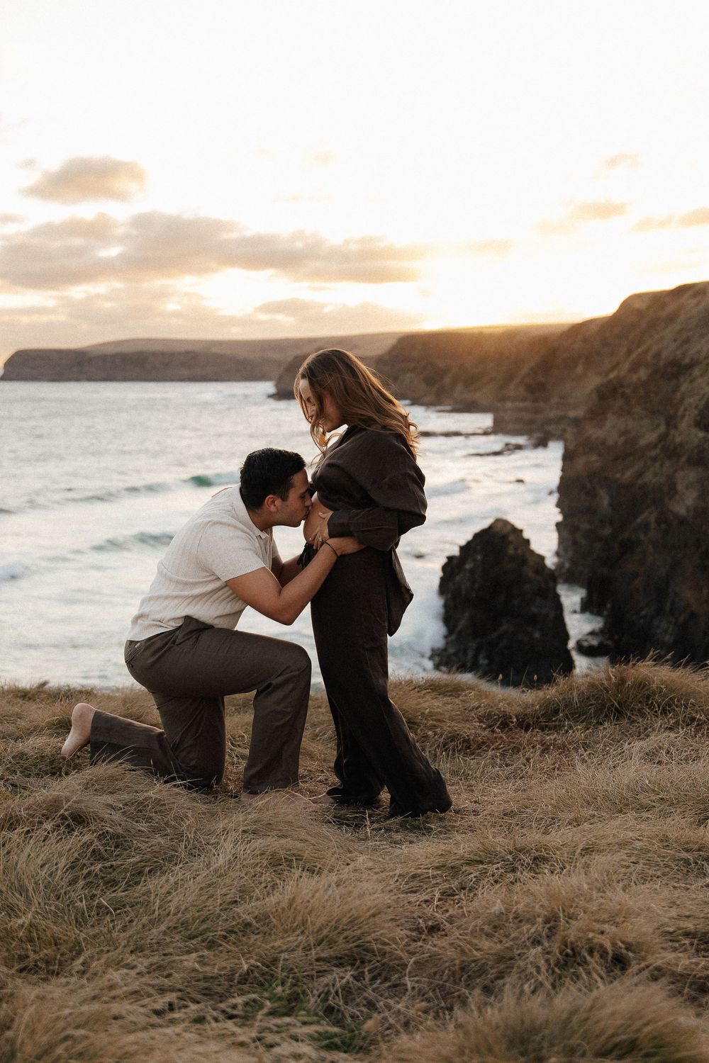 Couple sharing a romantic moment at sunset on a coastal cliff, man kneeling and kissing woman's hands by the ocean.