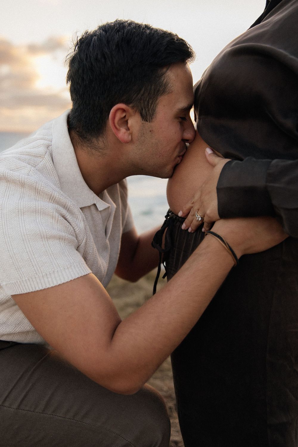 Man tenderly kissing pregnant woman's belly outdoors at sunset, with ocean in the background.