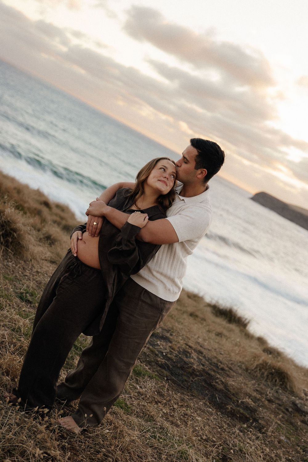Couple embracing at sunset on a coastal hillside, man kissing woman's cheek as ocean waves crash in background.
