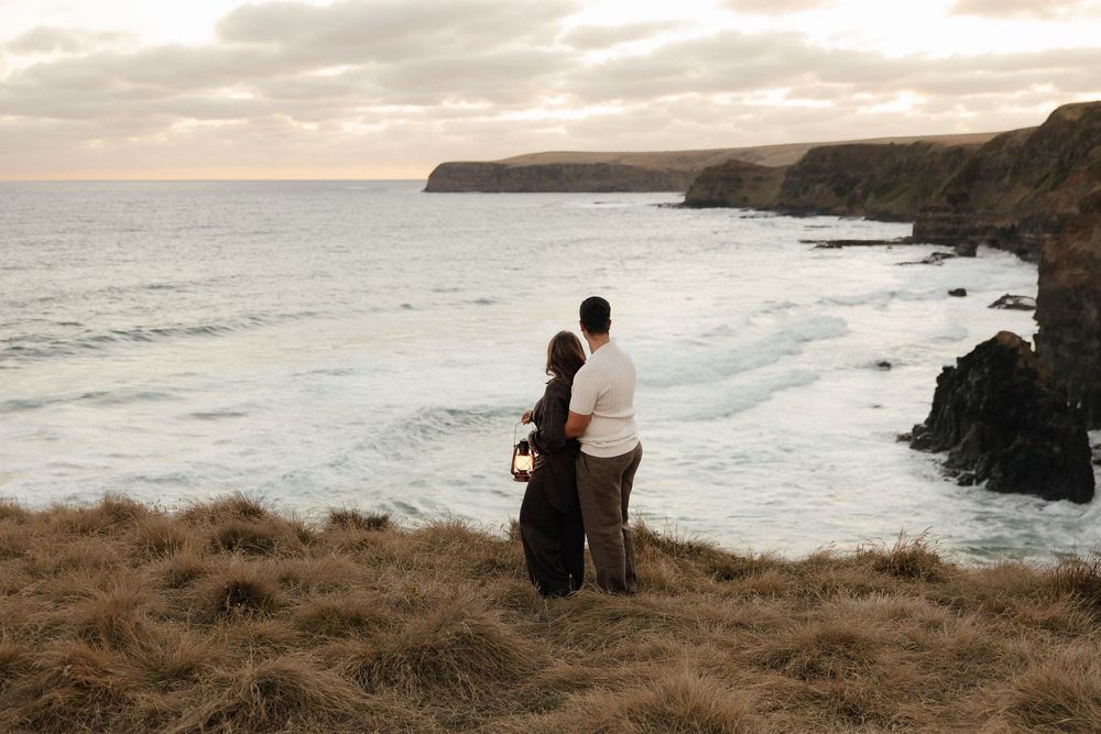 A couple embracing on a clifftop overlooking dramatic ocean waves and rocky coastline at sunset.