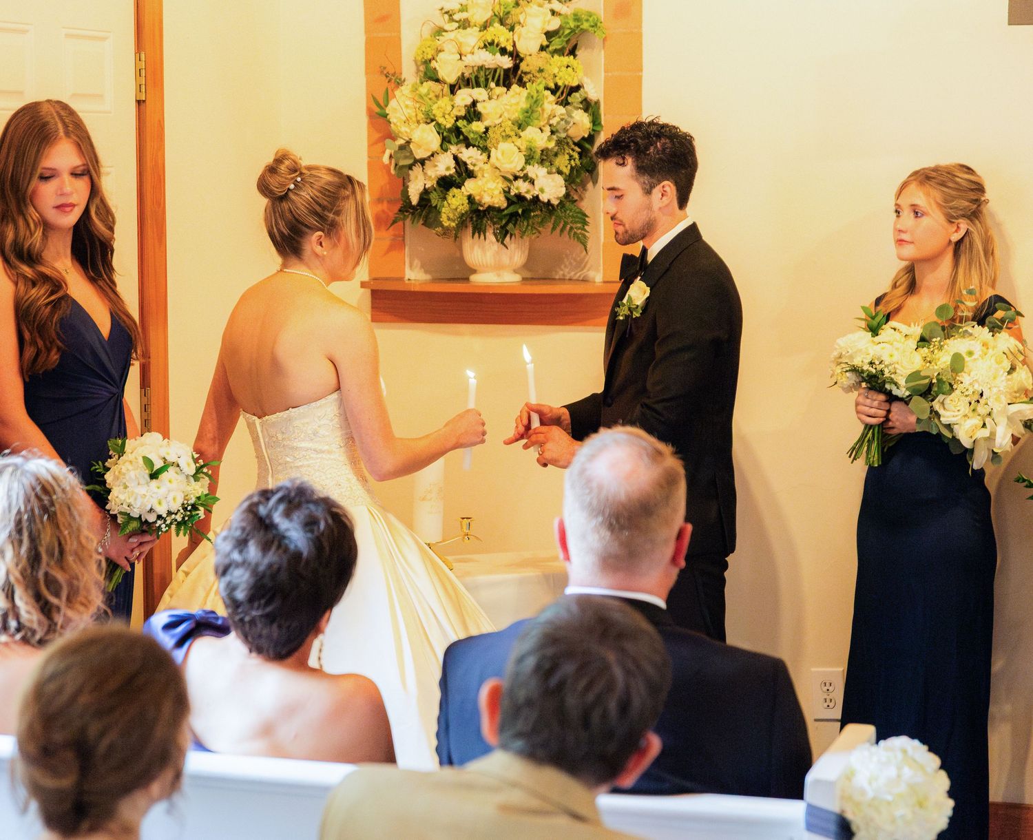 Bride and groom exchange vows at indoor wedding ceremony with bridesmaids in navy dresses holding white bouquets.